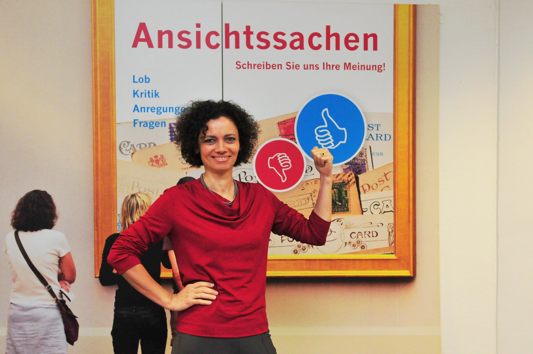 Birgit Bruckmann, Qualitätsmanagementbeauftragte der VHS. Die Mitarbeiterin steht vor einem Bild mit dem Titel Ansichtssachen. Dort können Teilnehmende Lob, Kritik, Anregungen und Fragen äußern. 